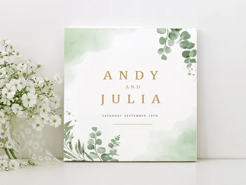 Elegant foam board sign with soft green watercolor foliage and gold typography, announcing the wedding of Andy and Julia, displayed next to delicate white flowers